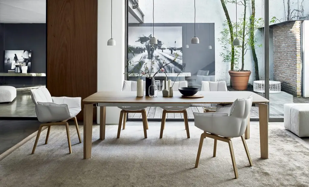 Wood dining table with white upholstered chairs beside a large window and courtyard tree