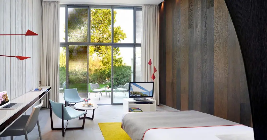 Hotel room with glass-walled area, wood TV console, yellow rug and bed with red trim