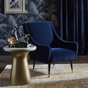 Navy blue velvet armchair with brass legs beside a round brass side table against botanical wallpaper