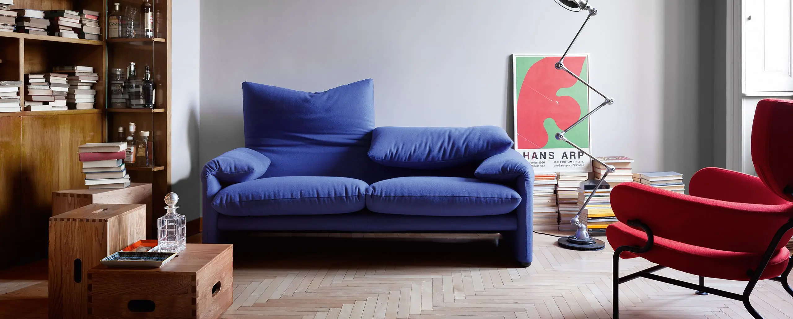Cassina Maralunga blue sofa beside a wooden bookshelf, red armchair and herringbone parquet floor
