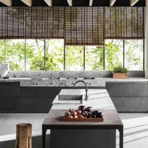 Concrete kitchen island with a stone countertop, bowl of fruit and bamboo blinds by a leafy window