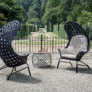 Two black wicker high-backed lounge chairs and a round side table on gravel facing a formal garden