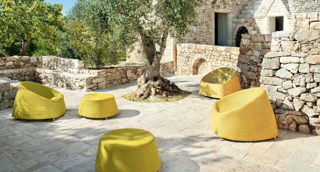 Mustard yellow quilted lounge chairs and poufs on a stone terrace beneath an olive tree at a rustic villa