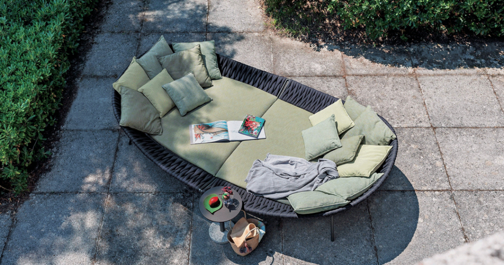 Aerial view of a round black wicker daybed with sage green cushions, a magazine and a small side table on a stone patio