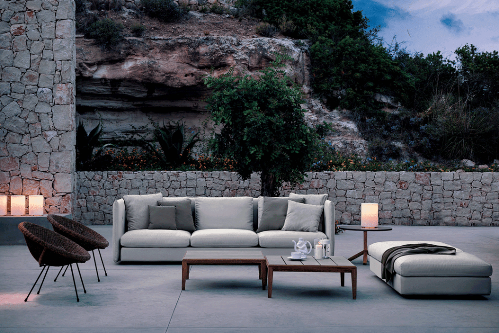 Evening patio with a stone wall, cream sofa, dark wicker chairs, wooden coffee tables and candles by a hillside