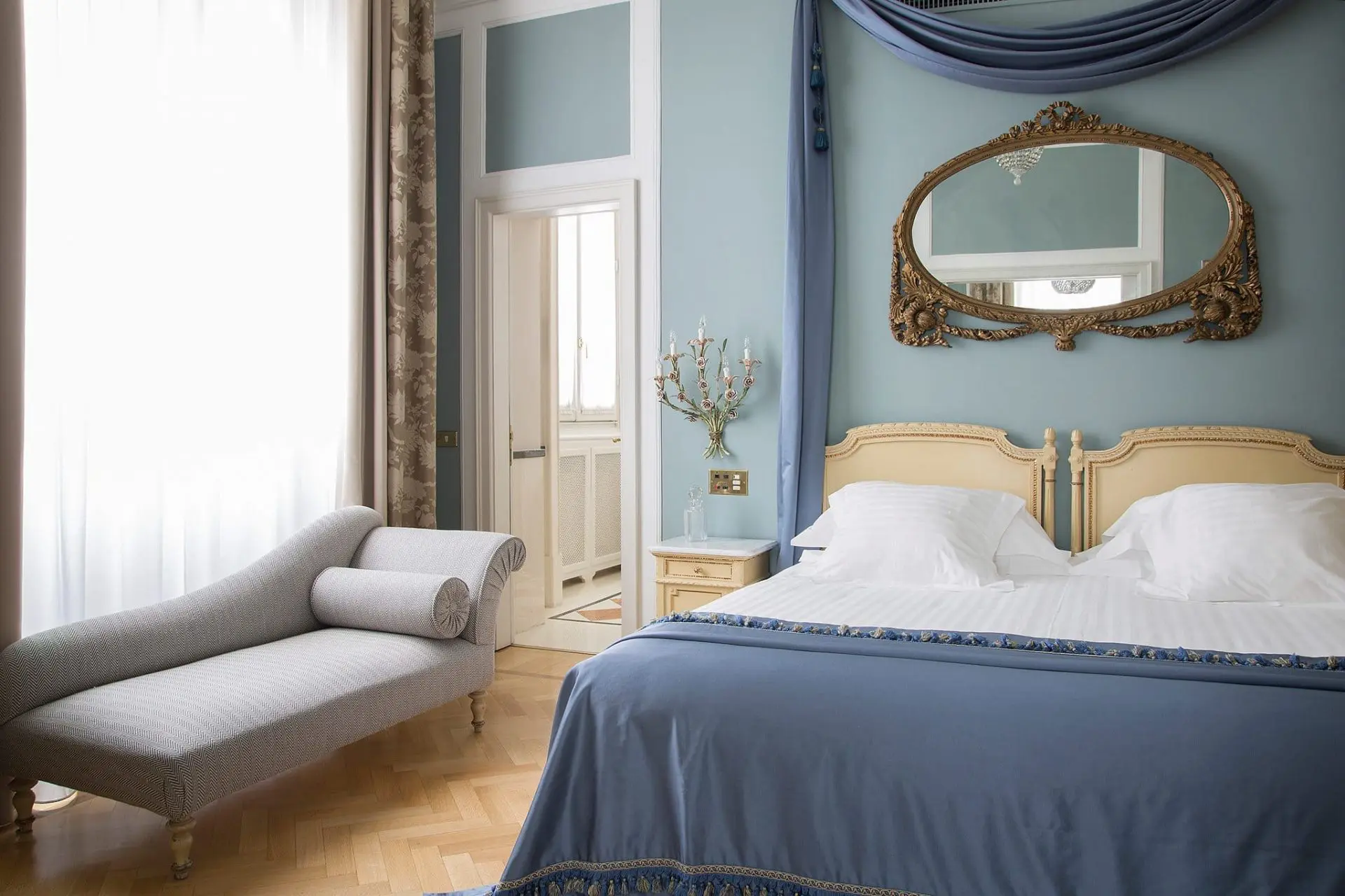 Blue hotel bedroom with an ornate gilt mirror, canopy drape and a chaise longue by the window