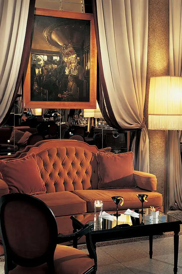 Hotel lounge corner with a tufted orange sofa and a framed painting under a fringed lamp