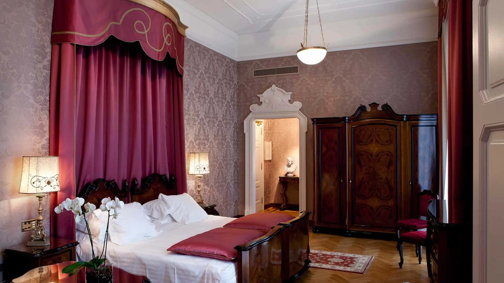 Ornate hotel bedroom with a red canopy bed, damask wallpaper and an antique wardrobe