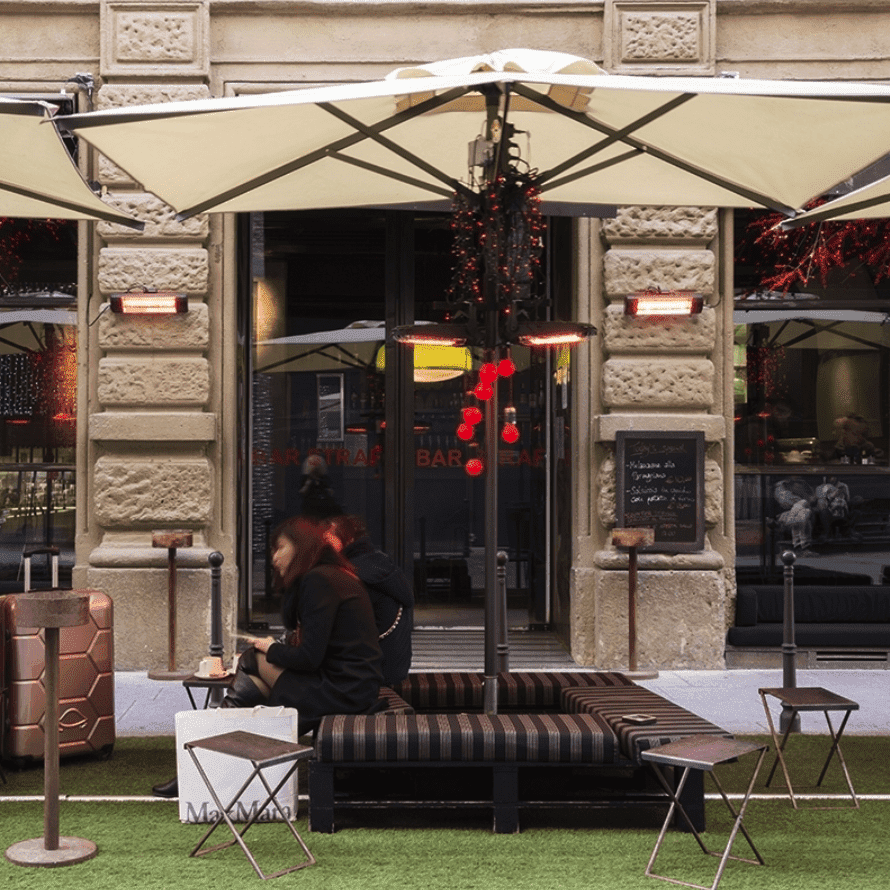 Outdoor café seating under a large umbrella on an artificial lawn beside a stone facade