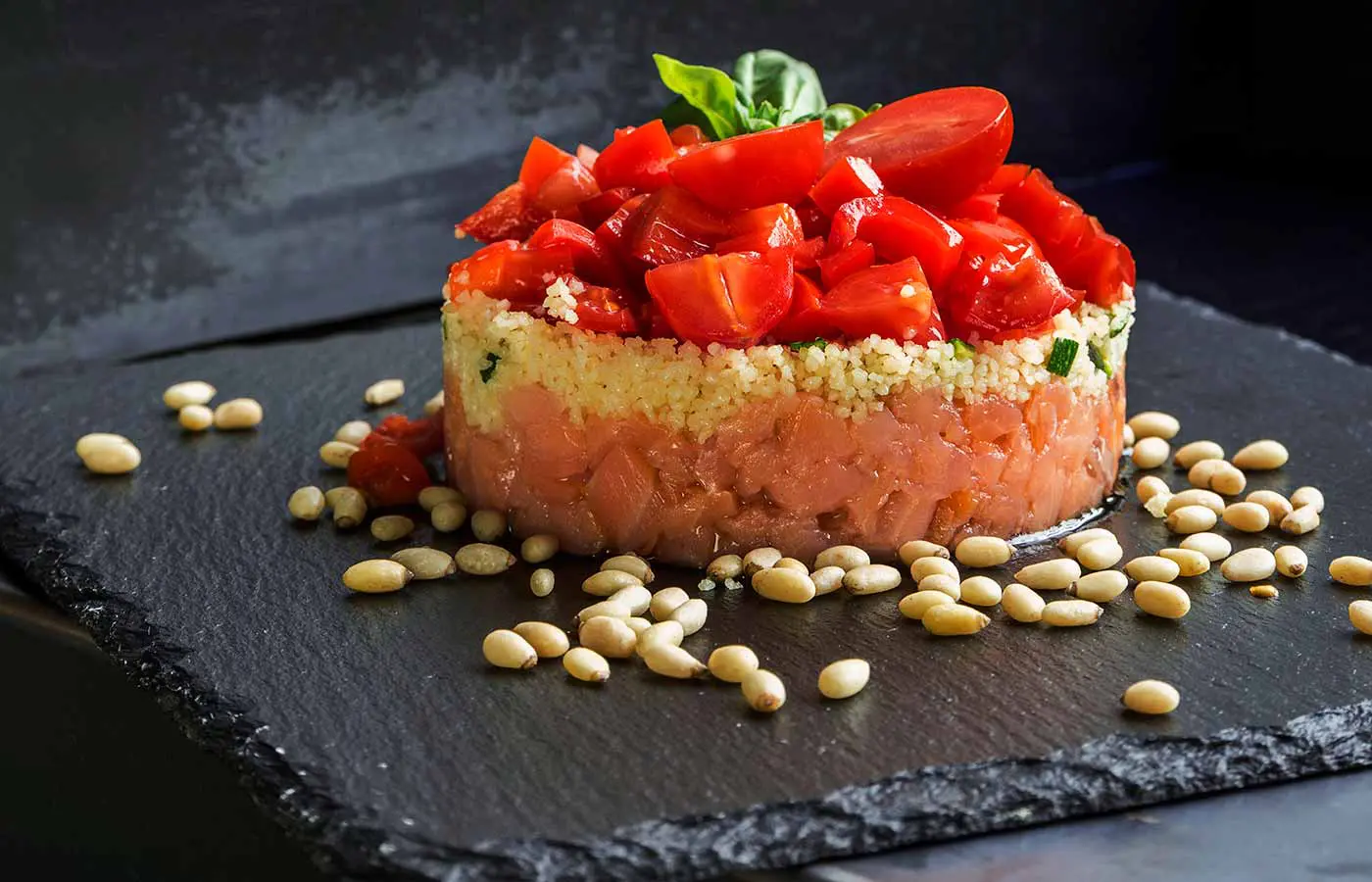 Salmon tartare topped with diced tomato and basil, plated on slate with pine nuts scattered around