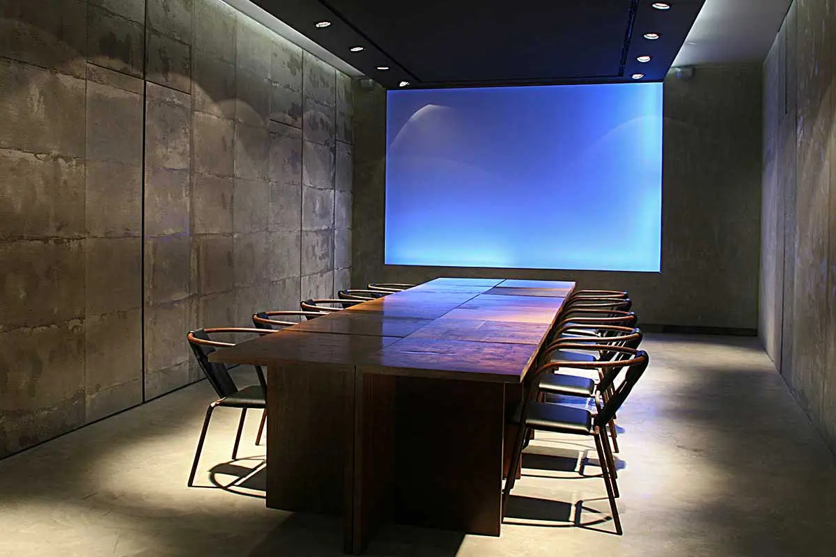 Dark conference room with a long wood table, chairs and a glowing blue screen