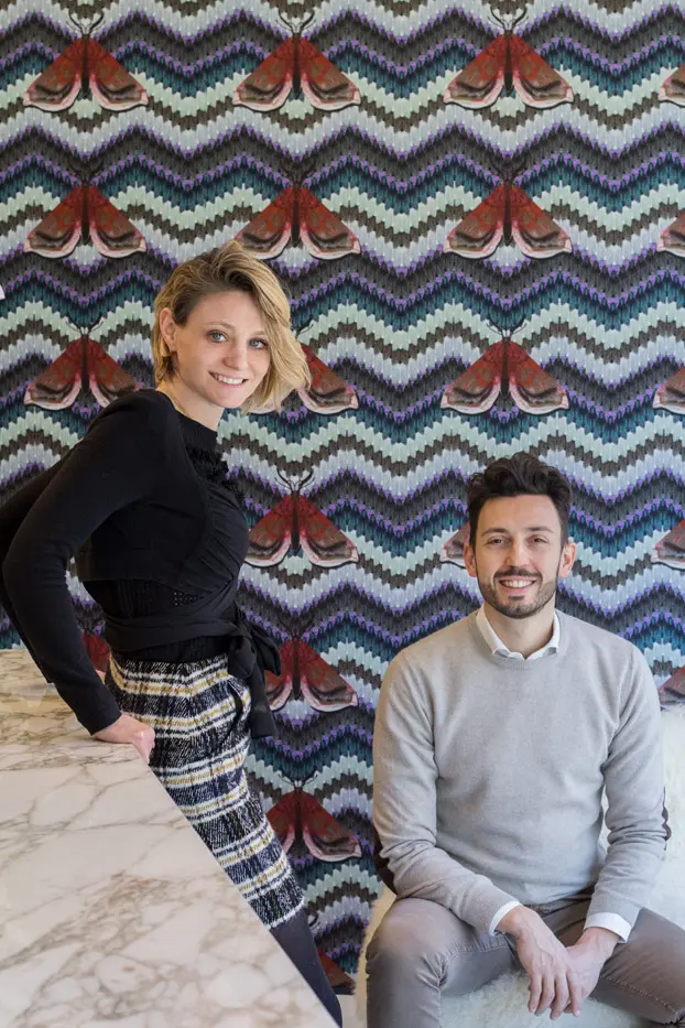 Man and woman posing for a portrait against moth-patterned wallpaper near a marble counter