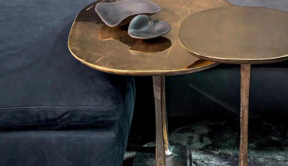 Organic brass side tables with sculptural glass objects beside a dark velvet sofa
