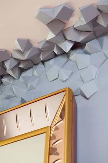 Faceted gray geometric wall panel surrounding a gold framed mirror
