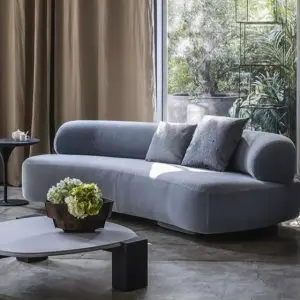 Grey velvet curved sofa with bolster armrests beside a round white marble coffee table