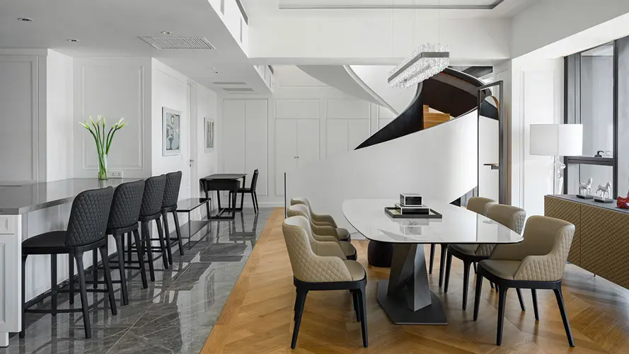 Dining area with a round marble table and beige chairs beside a curved staircase and chandelier