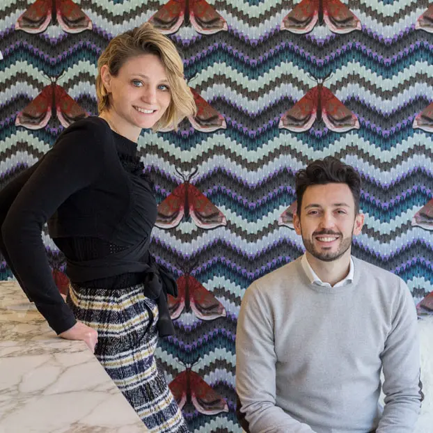 Man and woman posing for a portrait against moth-patterned wallpaper near a marble counter